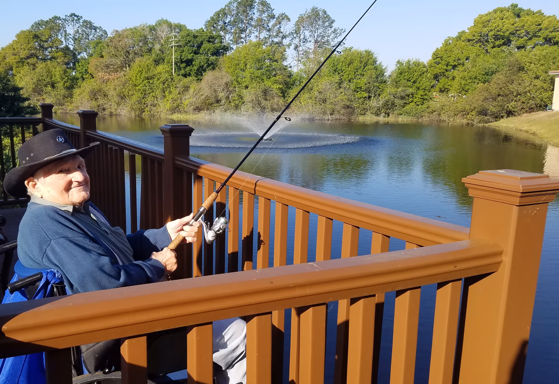 An elderly man wearing a black cowboy hat and blue jacket is sitting in a wheelchair on a wooden deck, fishing in a pond with a fountain in the background surrounded by trees and greenery.