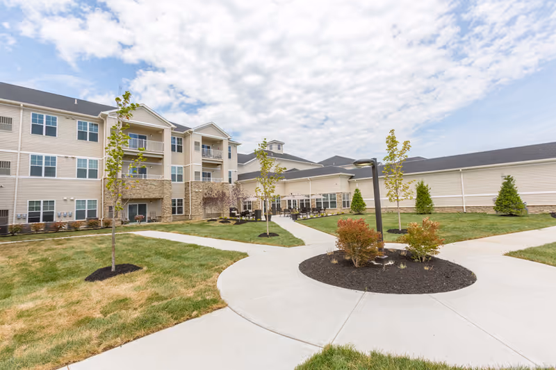 Outdoor courtyard area of a senior living facility with a three-story beige building in the background, young trees planted in mulched beds, green grass, concrete walkways, and outdoor seating with tables and chairs under a partly cloudy sky.