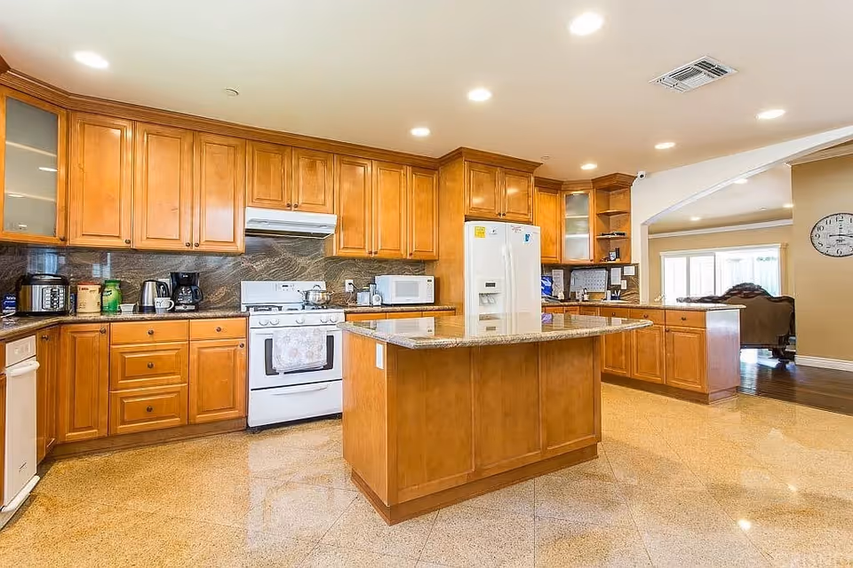 A spacious kitchen with wooden cabinets, a central island with a granite countertop, a white stove with a range hood, a white refrigerator, a microwave, and various small kitchen appliances on the countertops. The kitchen opens into a living area with a clock on the wall and large windows.