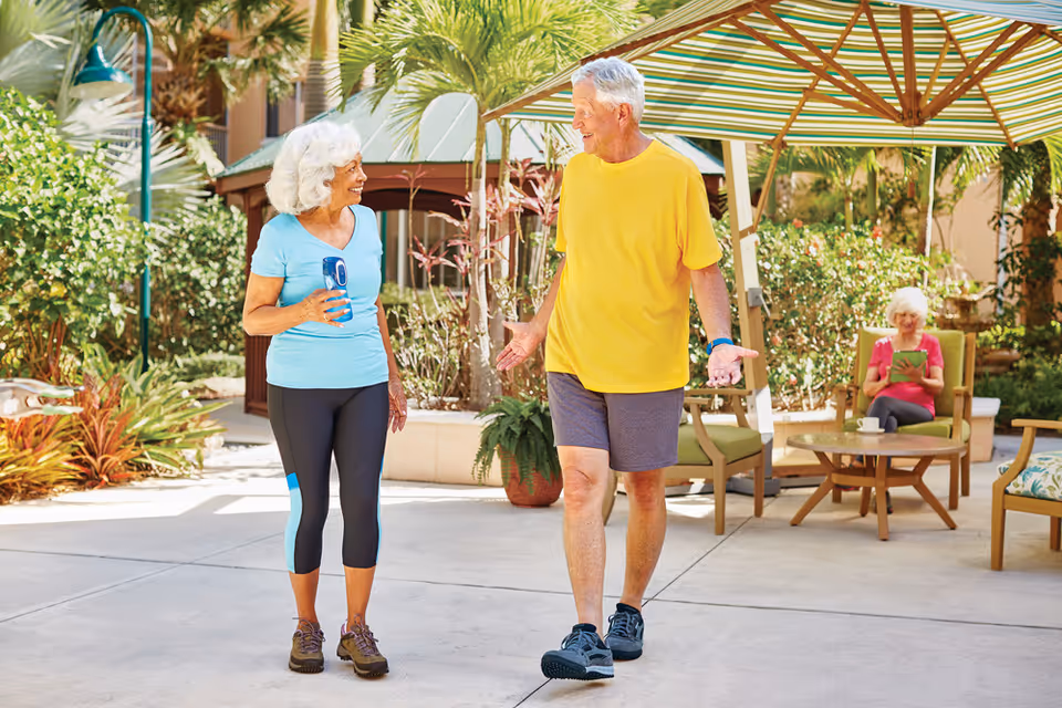 An elderly woman in a light blue shirt and black leggings holding a water bottle walks and talks with an elderly man in a yellow shirt and gray shorts under a large striped umbrella in a sunny outdoor garden area. In the background, another elderly woman in a pink shirt sits on a green cushioned chair reading a tablet.