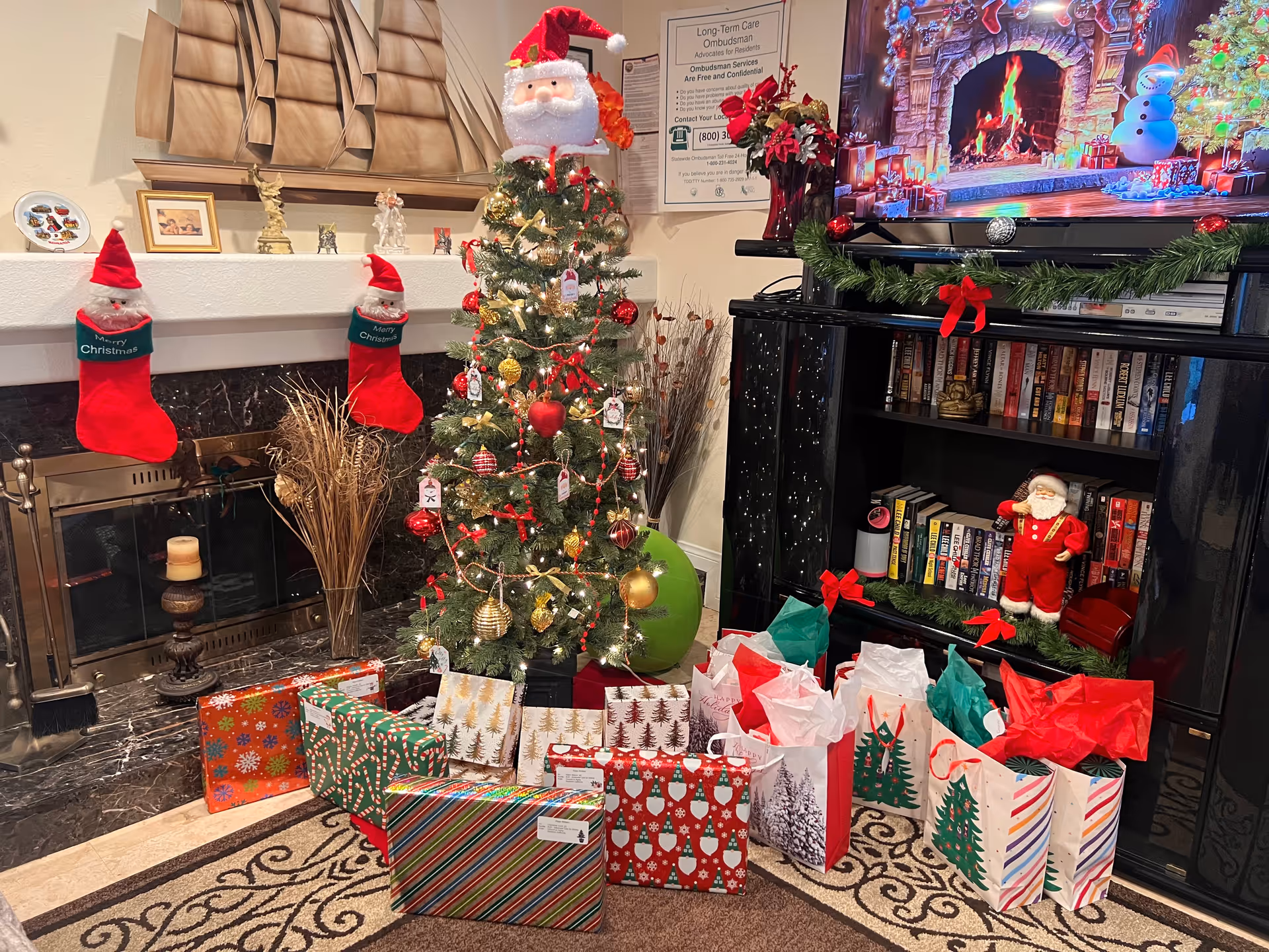 A cozy living room decorated for Christmas with a small Christmas tree adorned with ornaments and a Santa Claus hat on top. There are two red Christmas stockings hanging on the fireplace mantle, which also has decorative items including a model ship and framed pictures. In front of the tree, there are various wrapped gifts and gift bags. A TV above a black cabinet displays a festive fireplace scene with a snowman decoration on the right side of the cabinet. The cabinet shelves hold books and a small Santa figurine. The room has a warm and festive holiday atmosphere.