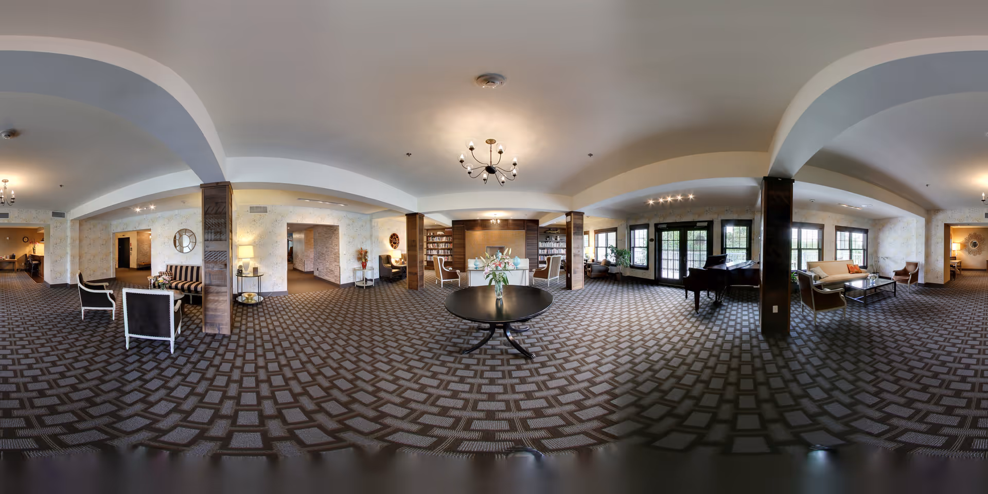 A spacious, well-lit living room area in The Residence at Otter Creek featuring patterned carpet flooring, multiple seating arrangements including sofas and armchairs, a grand piano near large windows, and a central round table with a vase of flowers. The room has arched ceiling details, several light fixtures, and decorative elements such as clocks and lamps on side tables.