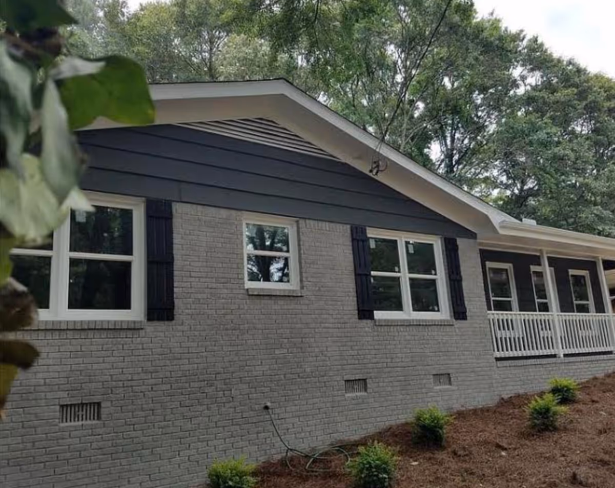 Exterior front of a single-story gray brick building with white-trim windows, black shutters, and a porch railing set among trees.