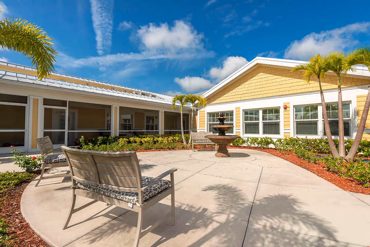 Outdoor courtyard area at The Brennity at Tradition Senior Living featuring a circular concrete patio with benches, a central water fountain, palm trees, and a yellow building with white trim under a blue sky with scattered clouds.