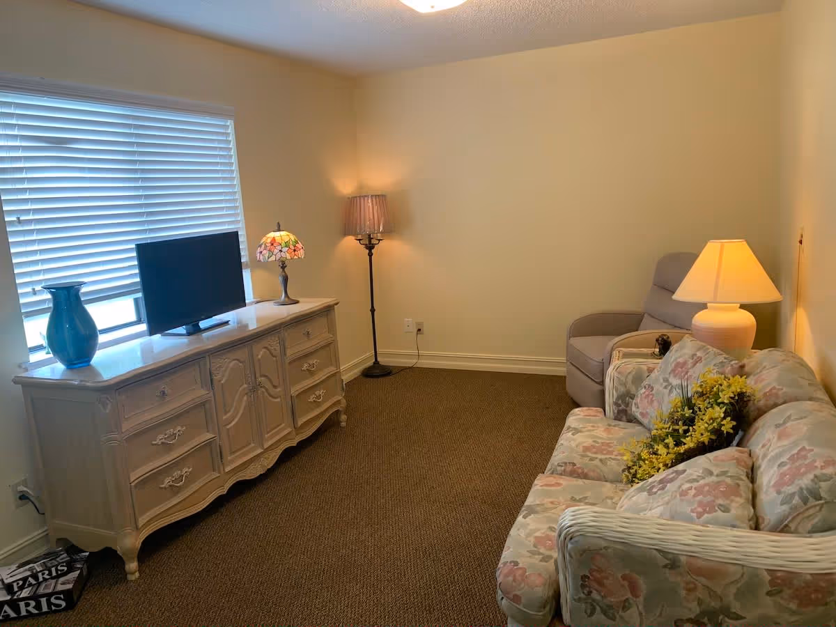 A cozy living room with a floral patterned sofa adorned with yellow flowers, a beige armchair, a side table with a lamp, a floor lamp in the corner, and a vintage-style dresser holding a flat-screen TV, a blue vase, and a stained glass lamp. The room has beige walls and a window with closed blinds.
