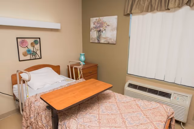 A small, simple bedroom in a care facility with a single bed featuring safety rails, a wooden overbed table, and a patterned bedspread. There is a wooden nightstand with a turquoise vase, a floral painting on the wall, and a window with vertical blinds and a valance. An air conditioning unit is installed below the window.