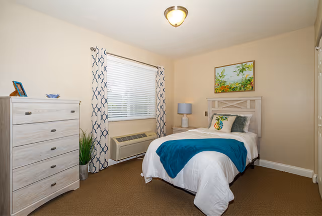 A cozy bedroom with a single bed dressed in white bedding and a teal throw blanket. The bed has decorative pillows and a wooden headboard. Next to the bed is a nightstand with a lamp. A window with patterned curtains and blinds is on the wall behind the bed. A light-colored wooden dresser with drawers is on the left side of the room, along with a potted plant. A colorful painting hangs above the bed on a beige wall.