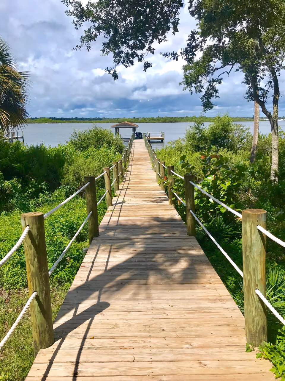 A wooden boardwalk with rope railings on both sides leading to a small covered dock over a body of water, surrounded by lush green vegetation and trees under a partly cloudy sky.
