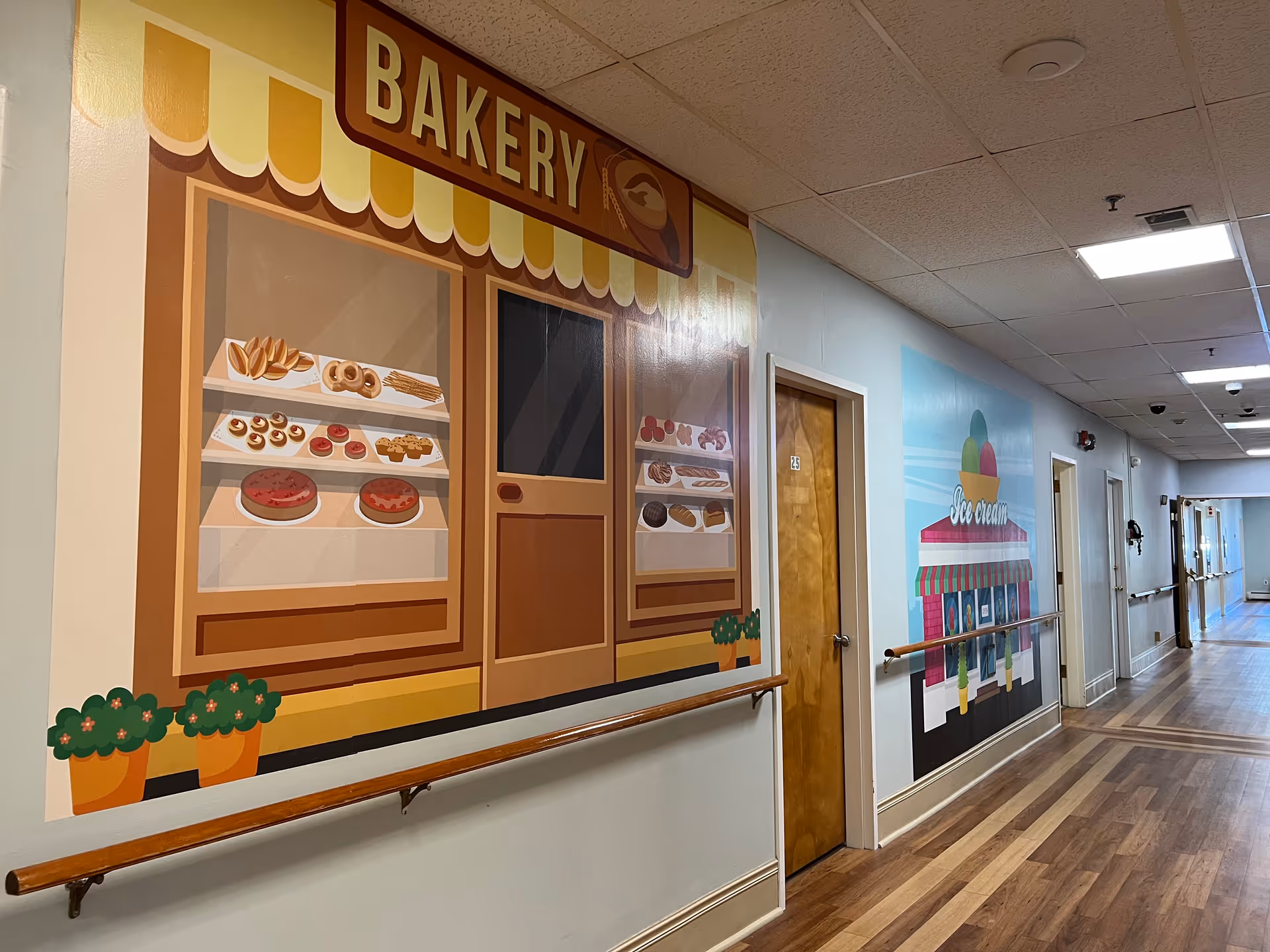 A hallway in a senior living community with wooden floors and handrails along the walls. The walls are decorated with colorful murals depicting a bakery storefront with pastries and an ice cream shop. Several closed wooden doors line the hallway under a drop ceiling with fluorescent lighting.
