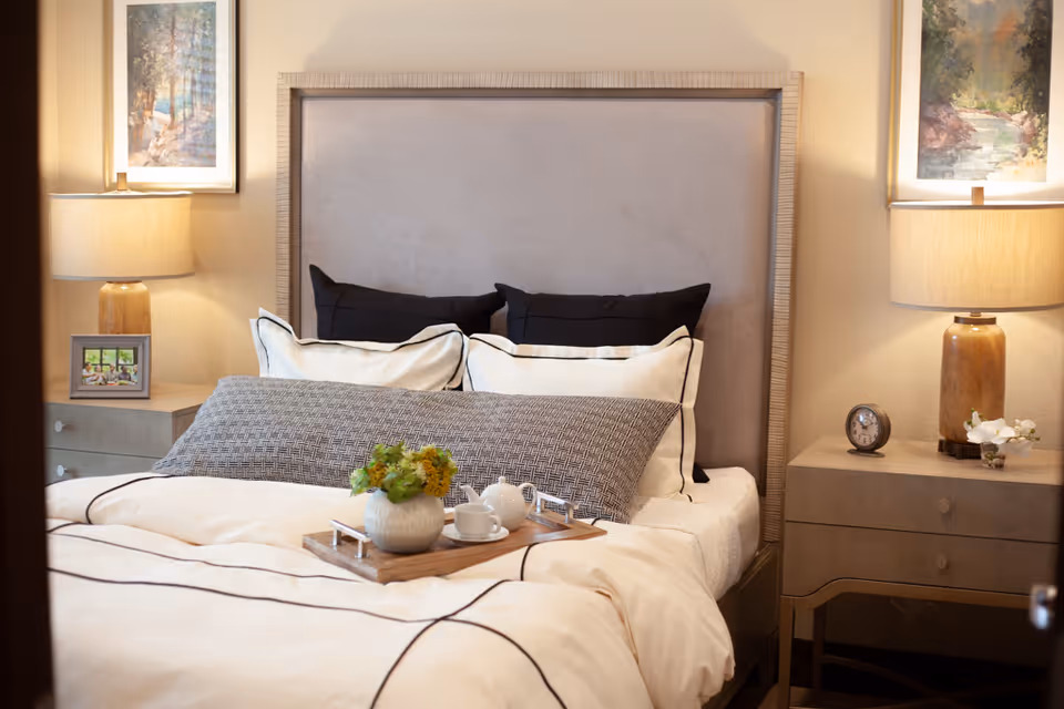 A neatly made bed with a beige upholstered headboard, white and black pillows, and a decorative gray pillow. A wooden tray with a small vase of flowers, a teapot, and a cup is placed on the bed. On each side of the bed are matching nightstands with table lamps, a small clock, a framed photo, and a small flower arrangement. Two framed landscape paintings hang on the wall above each nightstand.