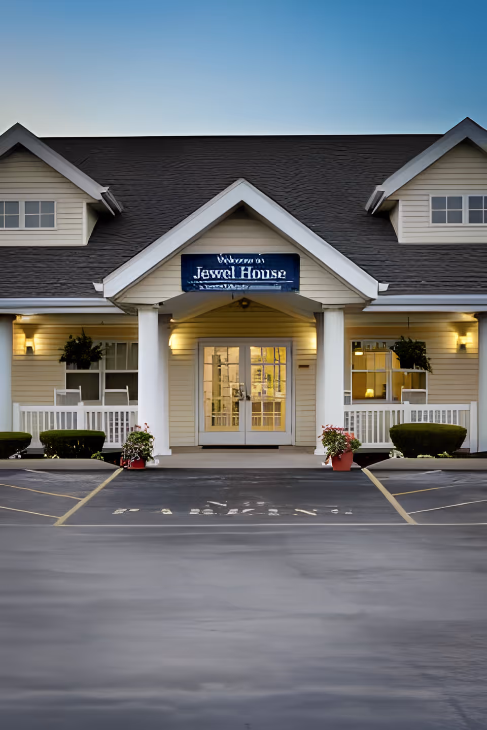 Front exterior view of Jewel Place Senior Living building with a peaked roof, white columns, and a porch with benches and potted plants on either side of the entrance. The parking lot is visible in the foreground.