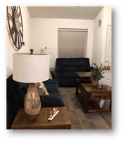 Small living room with dark upholstered sofas, wooden coffee and end tables topped with a lamp and decorative items, a large wall clock, and a window with closed blinds.