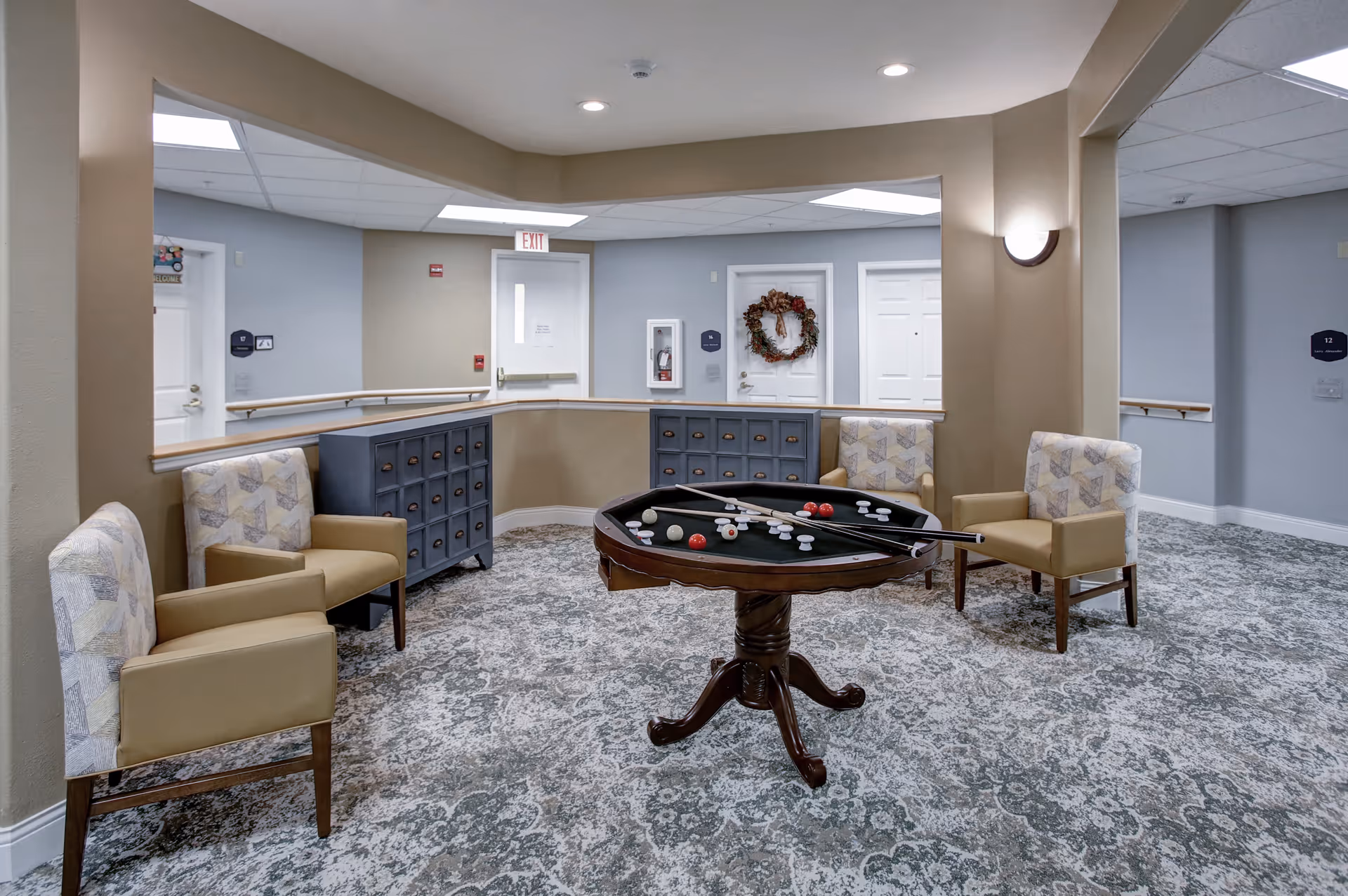 A cozy common area in a senior living facility featuring a round pool table with cues and balls, surrounded by four cushioned armchairs. The room has patterned carpet flooring, beige and light blue walls, and several white doors, one decorated with a wreath. There are two blue cabinets with multiple drawers along the walls and ceiling lights providing illumination.