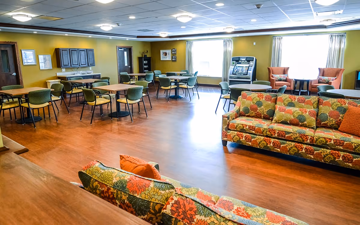 A spacious common area with multiple tables and chairs arranged for dining or socializing. The room features two floral-patterned sofas with orange accent pillows, two orange armchairs near large windows with curtains, and a jukebox against the far wall. The walls are painted yellow-green and the floor is wood.