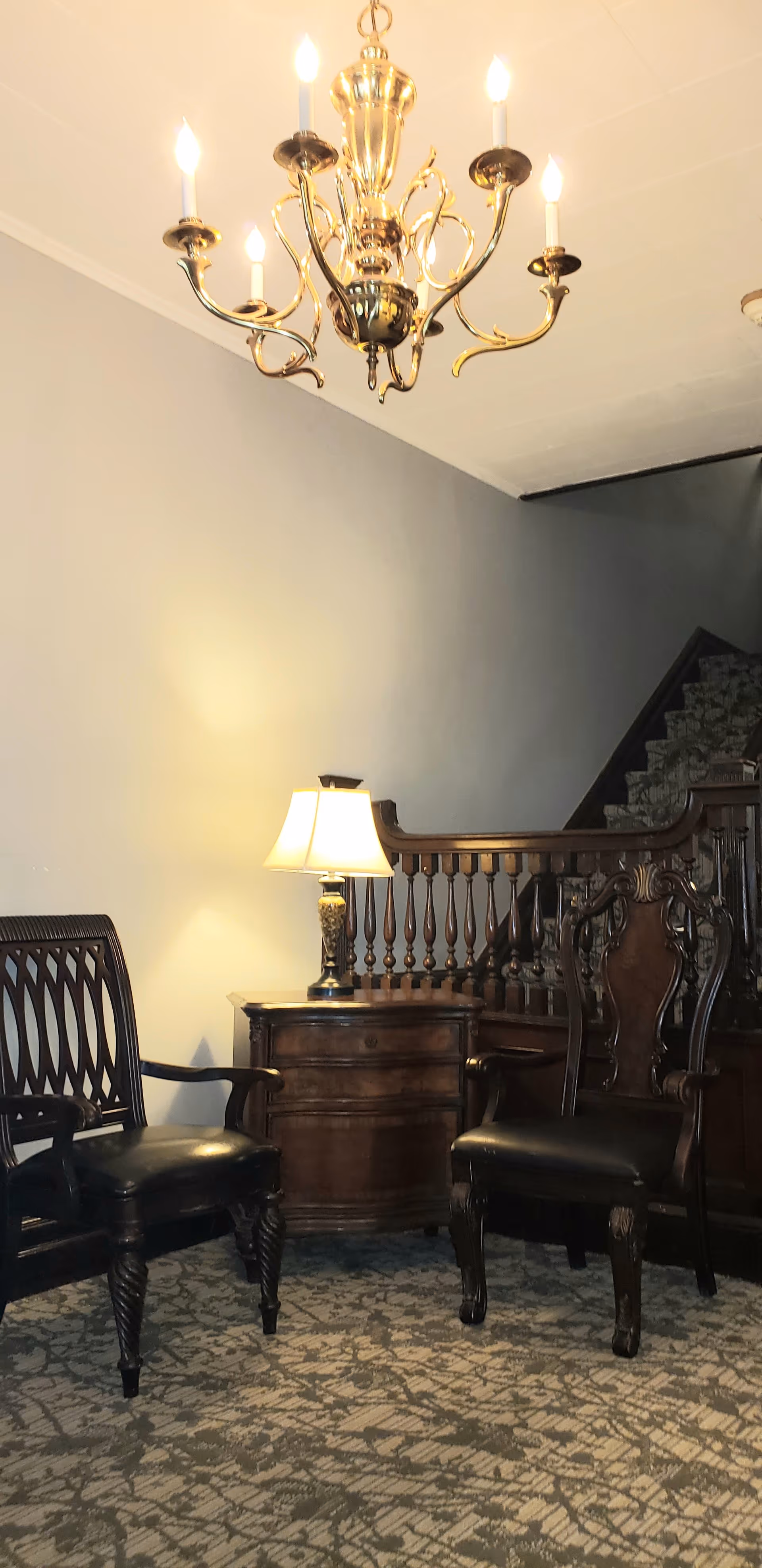 A cozy interior corner featuring two dark wooden chairs with black cushions, a wooden side table with a lit table lamp, a decorative wooden railing, a carpeted floor with a patterned design, and a brass chandelier with six lit bulbs hanging from the ceiling.