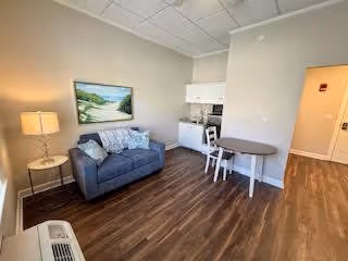 A small living area with a gray loveseat, a side table with a lamp, a round dining table with two chairs, and a kitchenette with white cabinets. The room has wood flooring and neutral-colored walls, with a landscape painting hanging above the loveseat.
