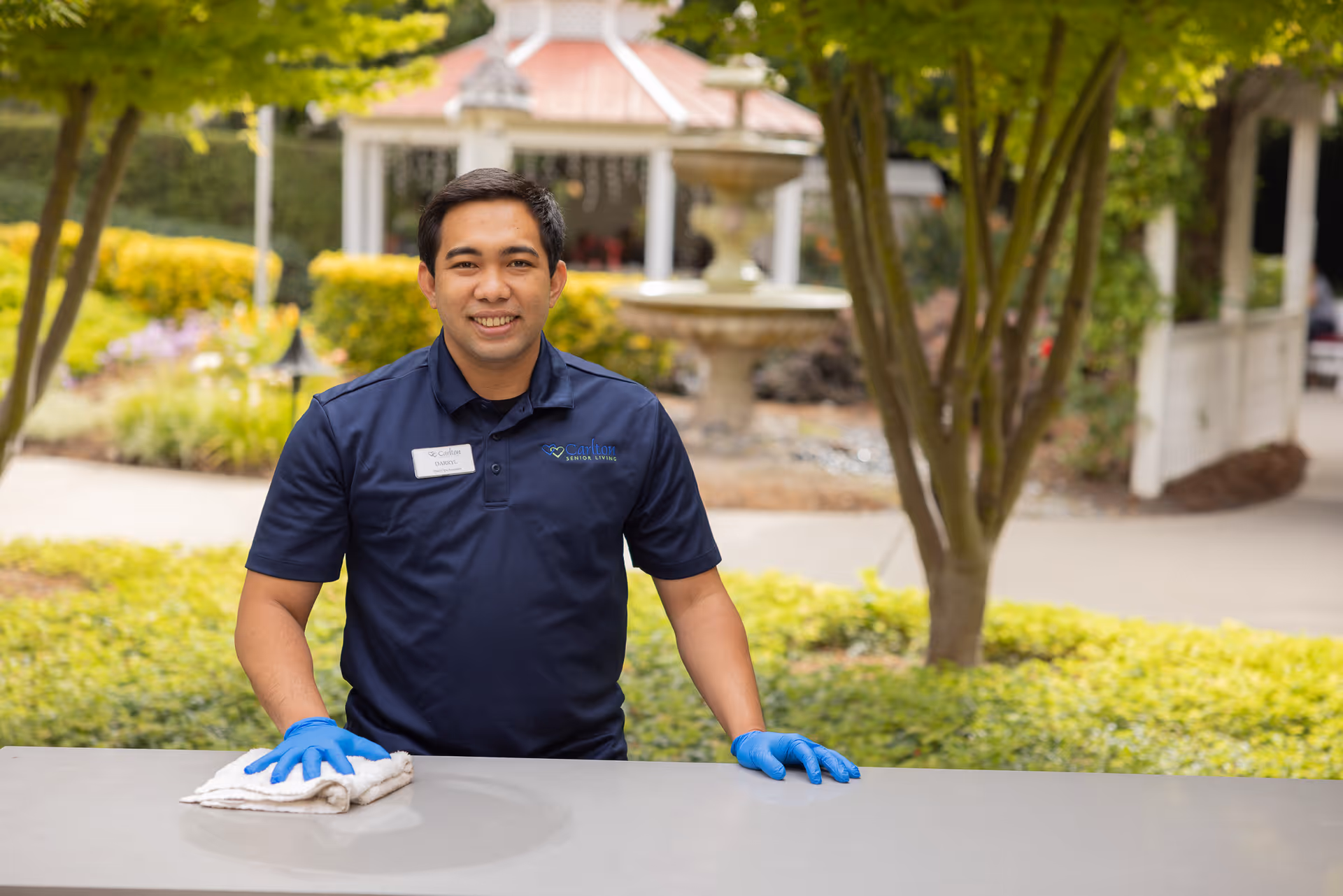 A man wearing a navy blue polo shirt with a Carlton Senior Living logo and blue gloves is cleaning an outdoor surface with a white cloth. Behind him is a garden area with green bushes, trees, a fountain, and a gazebo.
