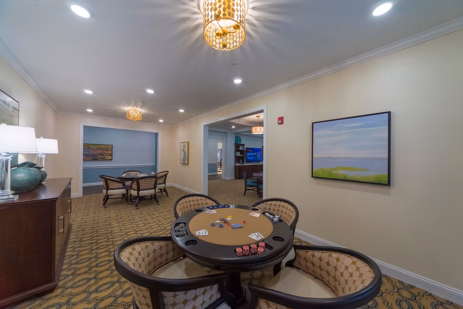 Interior view of a senior living facility common area with a poker table in the foreground surrounded by four cushioned chairs. The table has poker chips and playing cards on it. In the background, there are additional tables and chairs, wall art, and warm lighting fixtures on the ceiling. The walls are painted in neutral tones with carpeted flooring.