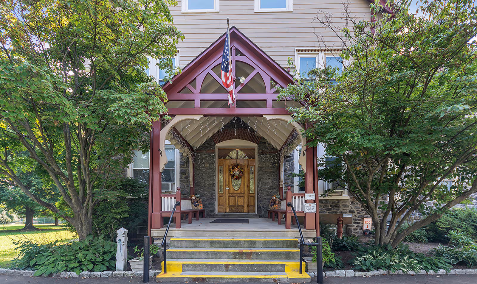 Front entrance of a building with a covered porch featuring a wooden door decorated with a wreath. The porch has a red and beige wooden frame with an American flag hanging above. There are steps leading up to the porch with yellow safety markings, flanked by black handrails. Trees and greenery surround the entrance.
