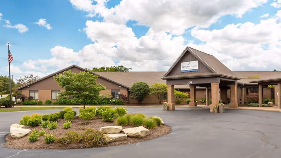 Exterior view of Brookdale Medina South senior living facility showing a single-story building with a covered entrance, landscaped roundabout with shrubs and rocks, an American flag on a flagpole, and a partly cloudy sky.