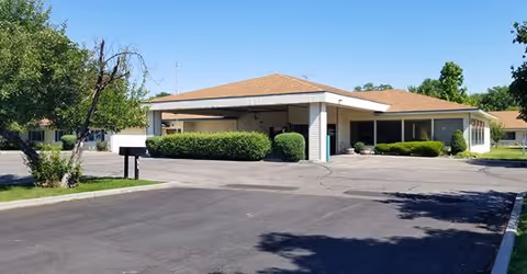 Single-story building with a covered entrance and surrounding greenery under a clear blue sky, with a paved driveway and parking area in front.