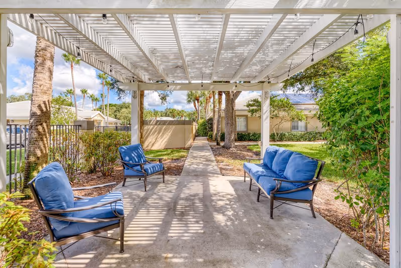 Outdoor seating area with blue cushioned chairs and a sofa under a white pergola, surrounded by greenery and trees, with a concrete pathway leading through the garden area.