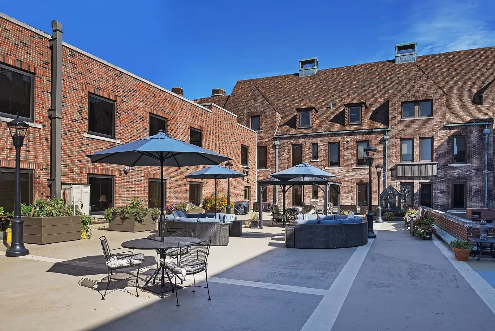 Outdoor courtyard area of a senior living facility with brick buildings surrounding it. The courtyard features several seating arrangements including round tables with chairs and umbrellas, as well as cushioned circular sofas under umbrellas. There are also planters with flowers and tall streetlamp-style lights. The sky is clear and blue.