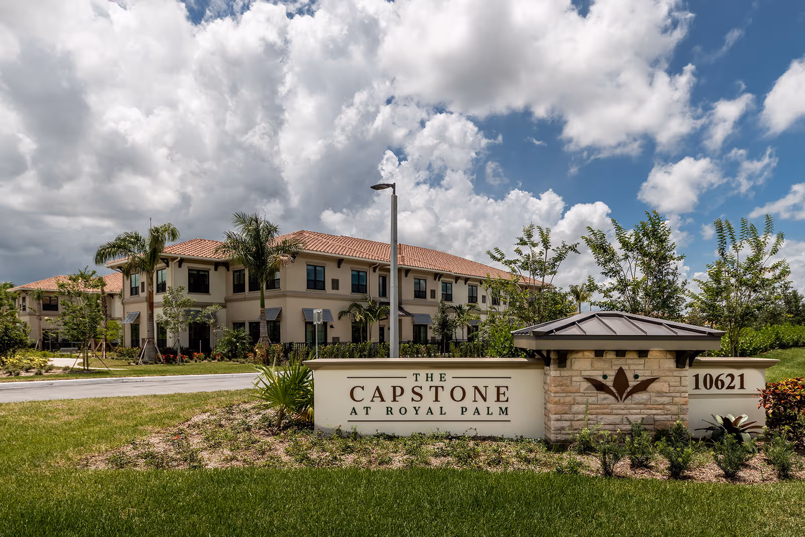 Entrance sign and landscaped lawn in front of The Capstone at Royal Palm two-story senior living building under a partly cloudy sky.