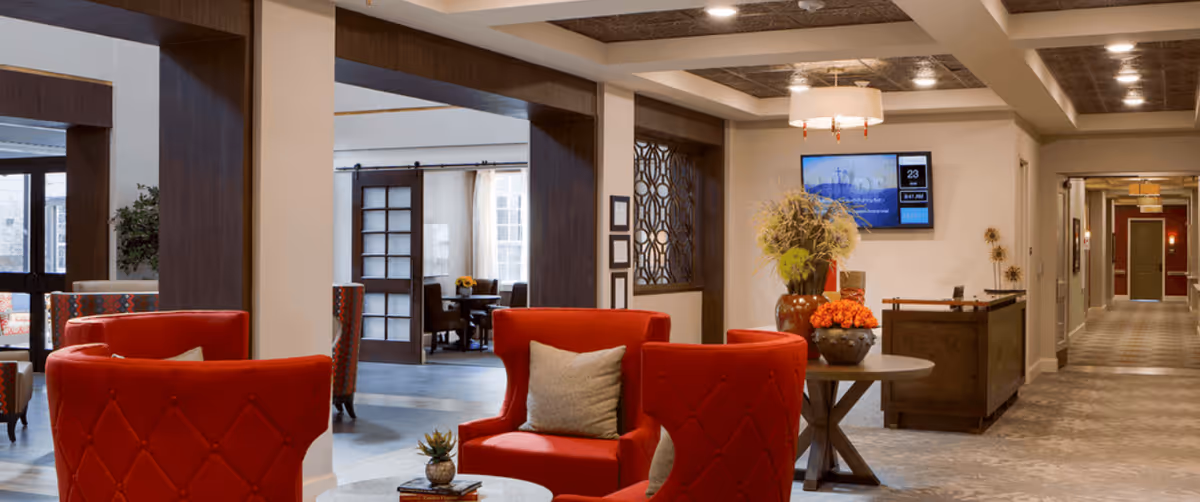 A spacious and well-lit senior living facility common area with red upholstered chairs arranged around small tables, decorative plants, and a hallway leading to other rooms. The area features warm lighting, wooden accents, and a wall-mounted screen displaying information.