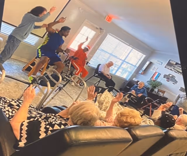 A group of elderly individuals seated in a common room participating in a seated exercise session led by two younger instructors. The room has large windows letting in natural light, and the elderly participants are raising their arms while seated on chairs and sofas.