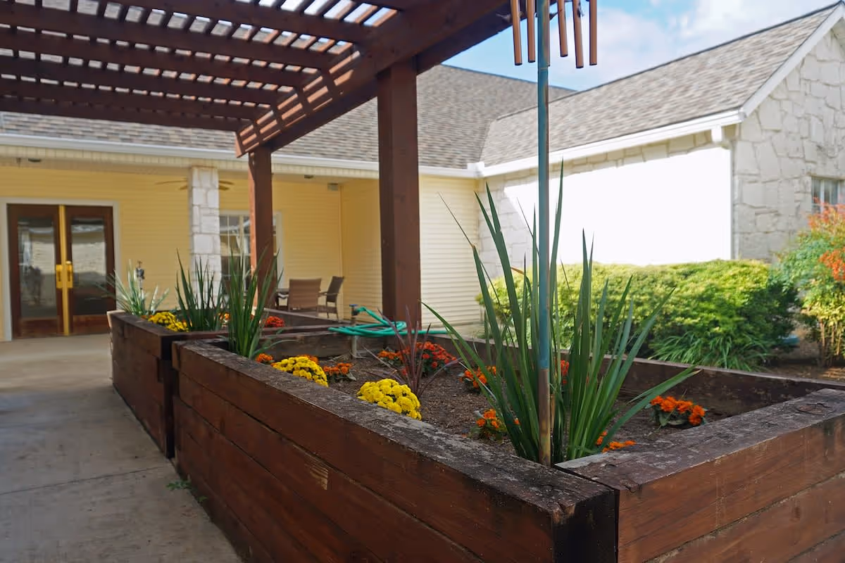 Outdoor garden area at New Haven Kerrville featuring raised wooden planter boxes with green plants and colorful flowers under a wooden pergola, with a building entrance and patio chairs in the background.