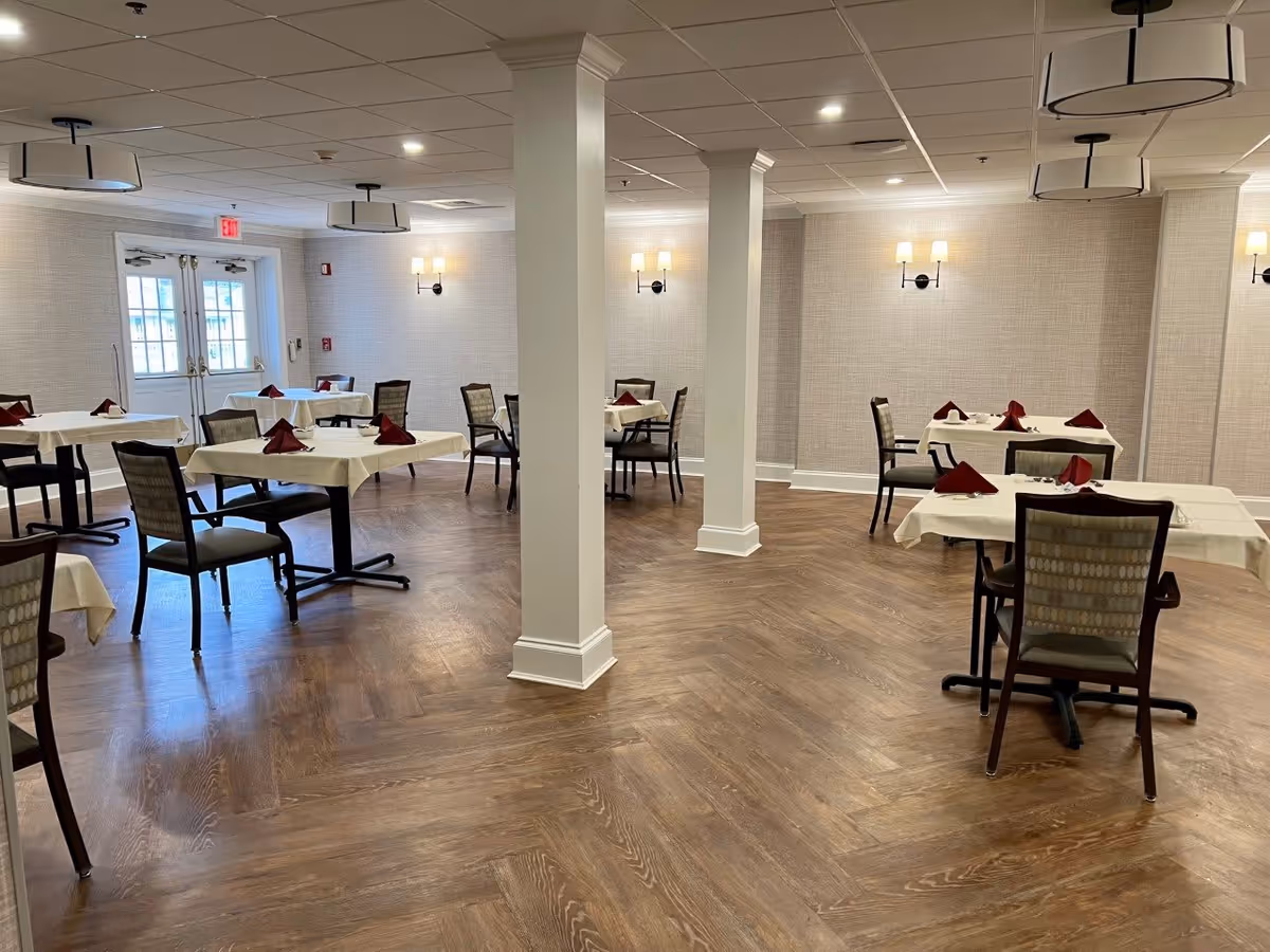 A dining room with several tables covered in white tablecloths and set with folded red napkins. The room has wooden flooring, beige textured walls, wall-mounted lights, and ceiling light fixtures. There are multiple chairs around the tables and double doors with windows at the far end of the room.