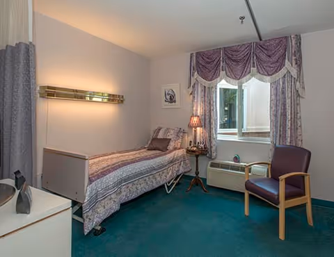 A small bedroom in a senior living facility featuring a single bed with patterned bedding, a bedside table with a lamp, a window with decorative curtains, a purple armchair, and a teal carpeted floor.