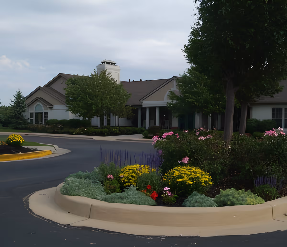 Front entrance of a one-story senior living building with a circular driveway and colorful landscaped flowerbeds.