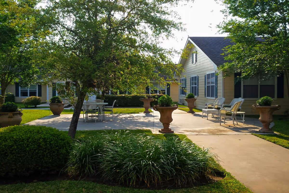 Outdoor patio area at Columbus Oaks Healthcare Community with a concrete seating area featuring white chairs and a table surrounded by large decorative planters. The patio is shaded by trees and surrounded by well-maintained green bushes and grass, with yellow buildings in the background.