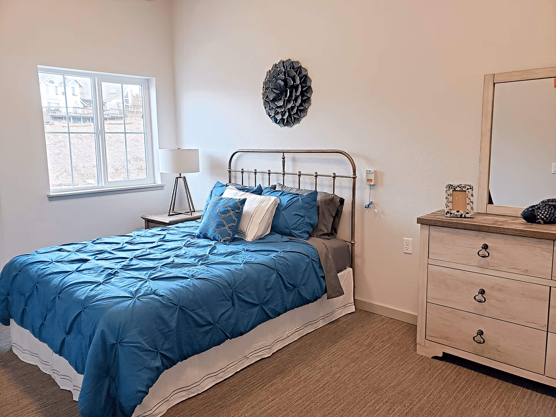 A bedroom with a metal bed frame, blue bedding, and multiple pillows. There is a wooden nightstand with a lamp next to the bed, a window with a view of houses outside, and a wooden dresser with a mirror and decorative items on top. A decorative wall piece hangs above the bed.