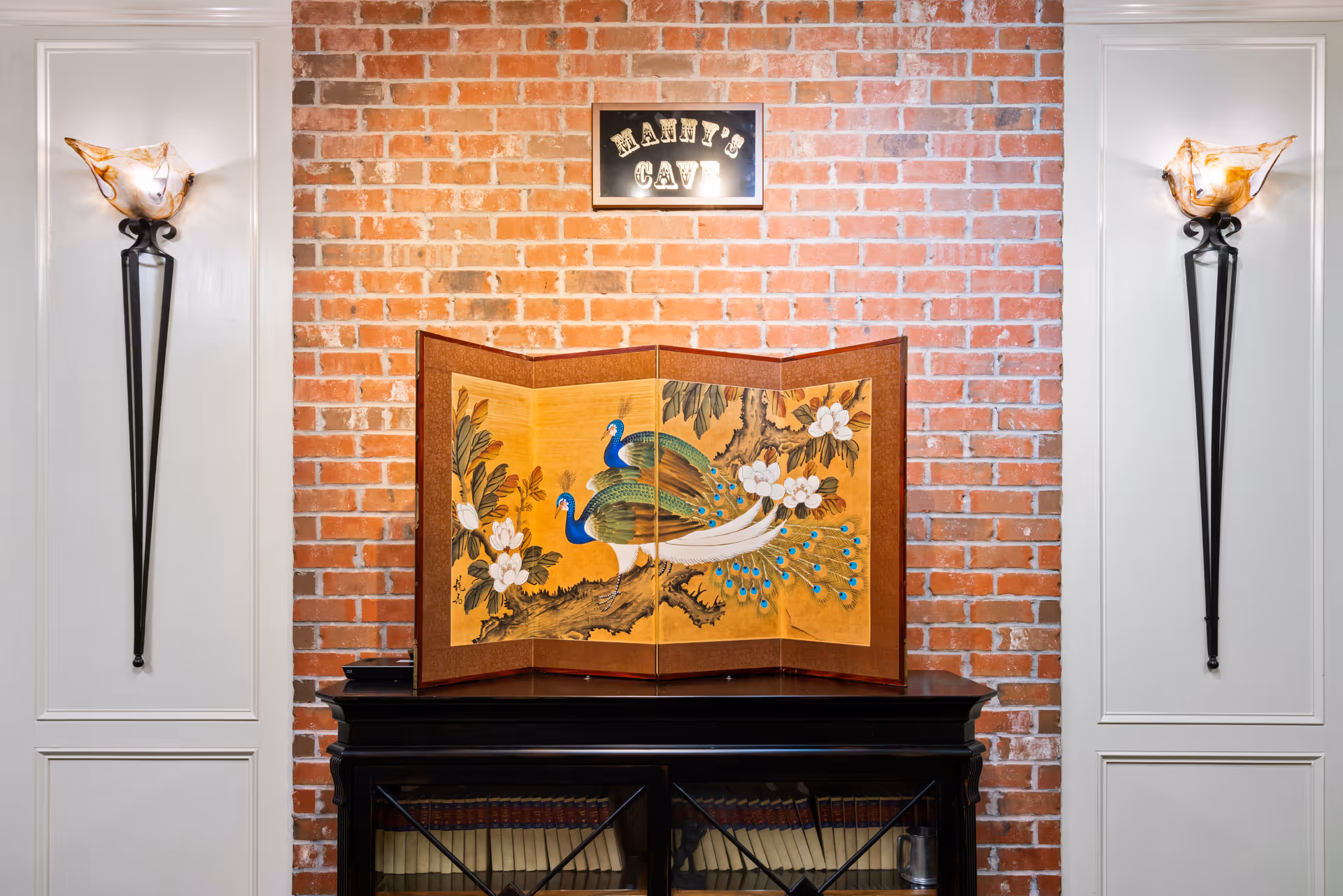 A decorative interior wall with exposed red brick and white paneling on either side. Two wall-mounted sconces with artistic glass shades are symmetrically placed on the white panels. A black cabinet with glass doors is positioned against the brick wall, topped with a folding screen featuring a painting of two peacocks on a tree branch with flowers. Above the screen, a small sign reads 'MANNY'S CAVE'.