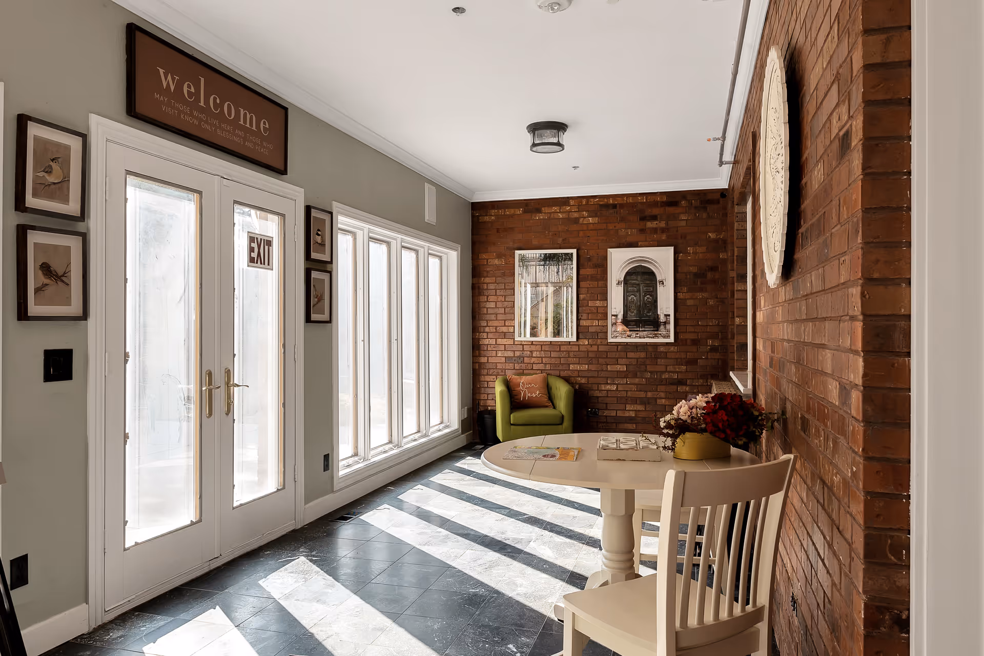 A bright interior room with large windows and glass doors letting in sunlight. The room features a brick wall with two framed pictures, a green armchair with a decorative pillow, a round white table with a flower arrangement, and a white chair. Above the doors is a wooden sign that says 'welcome'.