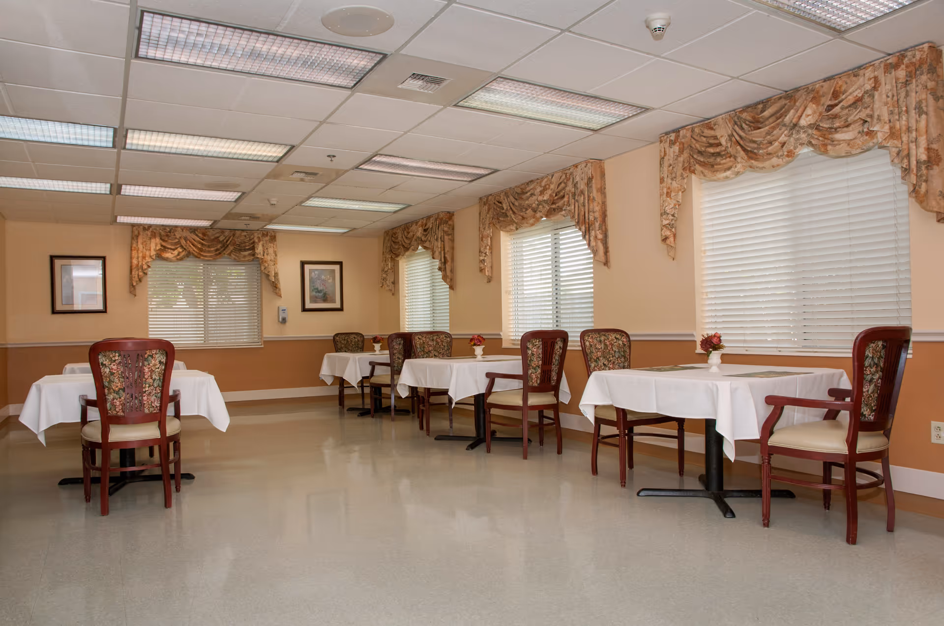 Small dining room with several tables set with white tablecloths and floral-upholstered chairs under curtained windows.