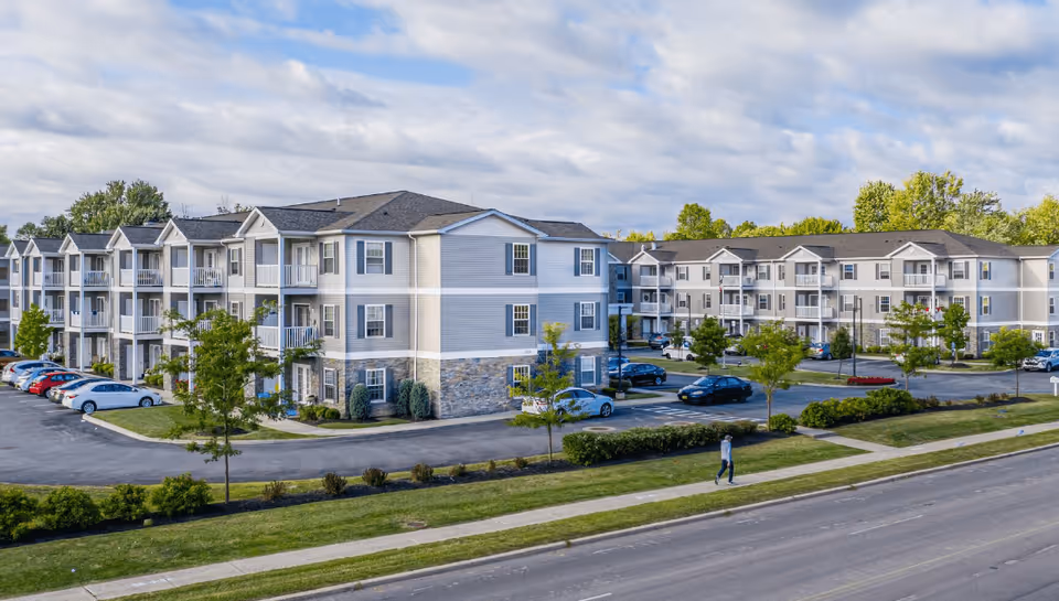 Exterior view of a multi-story senior living facility with beige siding and stone accents, surrounded by parking lots with cars and landscaped greenery under a partly cloudy sky. A person is walking on the sidewalk in front of the building.