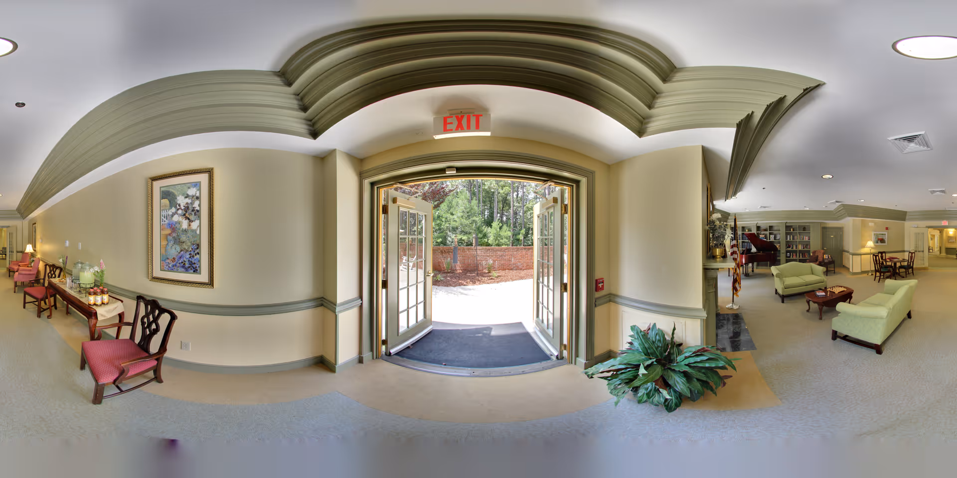 Bright senior living community lobby with double glass doors open to the outside, seating areas, plants, and decorative ceiling trim.