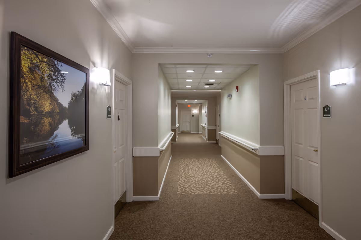 A well-lit hallway in a senior living facility with beige walls and carpeted floor. There are white doors on both sides of the hallway, each with room number signs. A framed picture of a river and trees hangs on the left wall. Handrails run along both sides of the hallway, and ceiling lights illuminate the space.