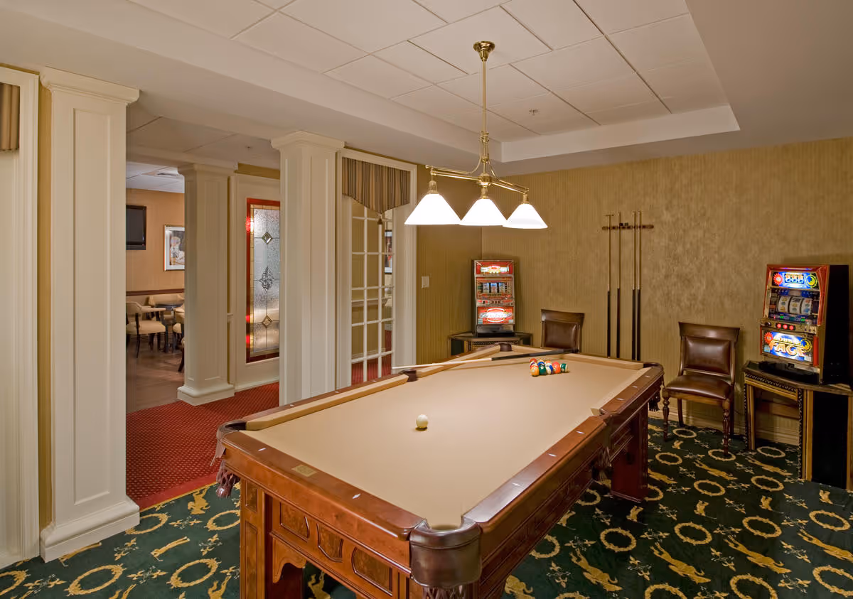 A carpeted game room with a billiards table, hanging light fixture, pool cues, chairs and slot machines.