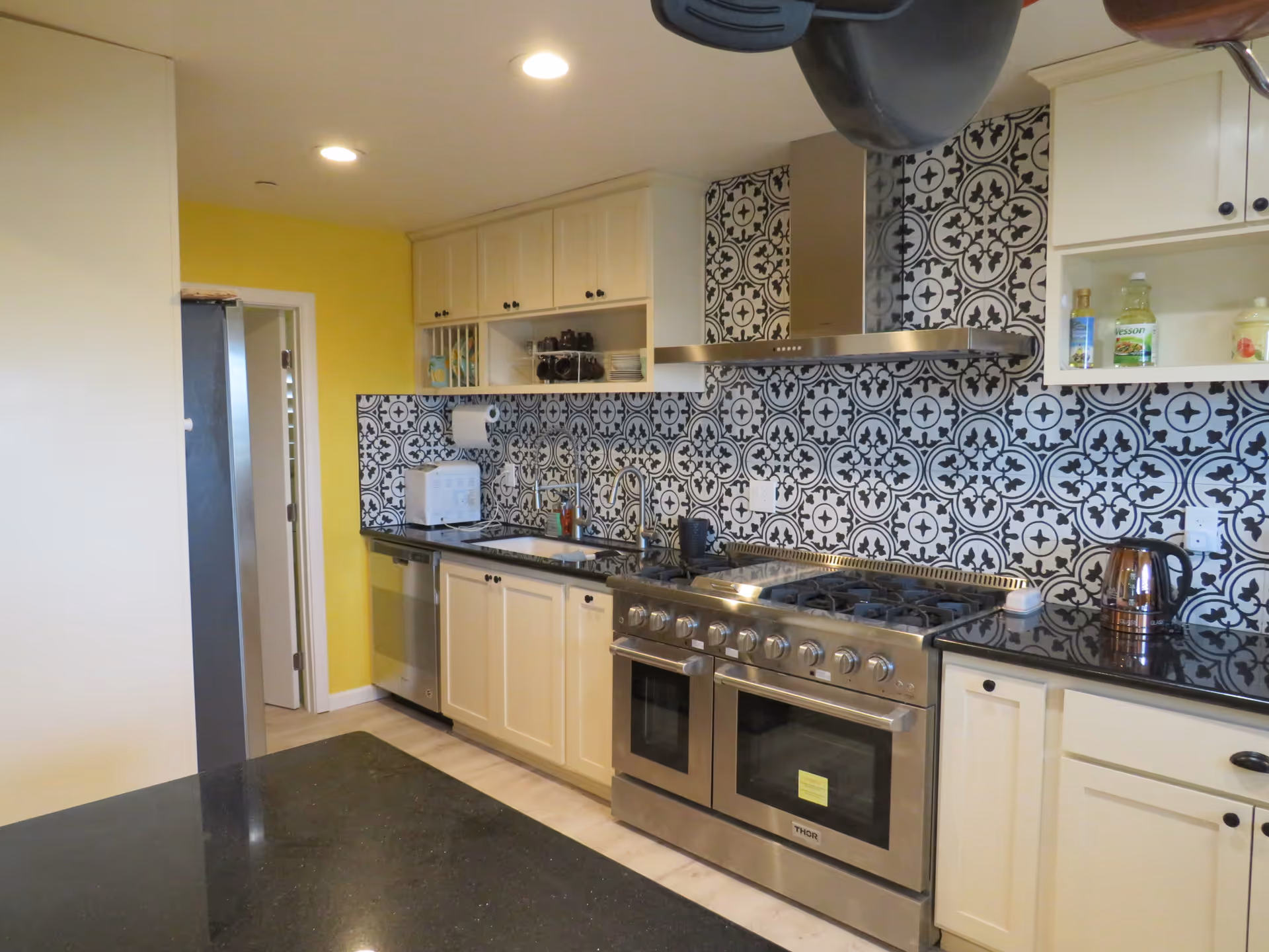 Modern kitchen with white cabinets, black and white patterned backsplash, stainless steel double oven and stove, black countertop, and yellow accent wall. There is a refrigerator, dishwasher, toaster, and various kitchen items on the counter.