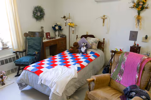 A cozy bedroom in a senior living facility with a single bed covered by a colorful quilt and a white blanket. The bed has stuffed animals and pillows on it. There is a wooden chair with a blue cushion near a window with light curtains. A wooden dresser with decorative items and flowers is against the wall. An armchair with a pink and green knitted blanket is also present. The walls have wreaths and religious decorations.