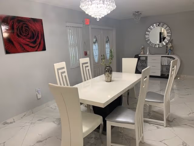 A modern dining room with a white rectangular table surrounded by six white chairs with silver cushions. A decorative vase with green plants is centered on the table. The room has white marble flooring, light gray walls, and a crystal chandelier hanging from the ceiling. On the wall to the left, there is a large red rose painting. At the back, there is a white sideboard with decorative items and a round mirror above it. Double doors with oval glass panels are visible in the background.