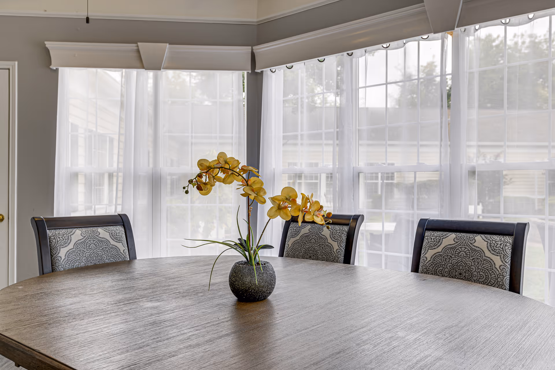 A round wooden dining table with a small vase holding yellow orchids in the center. Three upholstered chairs with patterned backs surround the table. Large windows with sheer white curtains allow natural light to fill the room.