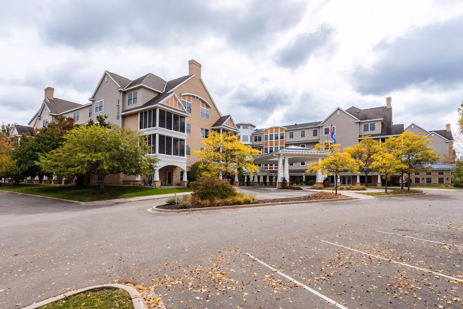 Exterior view of The Commons on Marice senior living facility showing a large multi-story building with beige and gray siding, multiple windows, and a covered entrance. The surrounding area includes trees with autumn foliage and an empty parking lot under a cloudy sky.