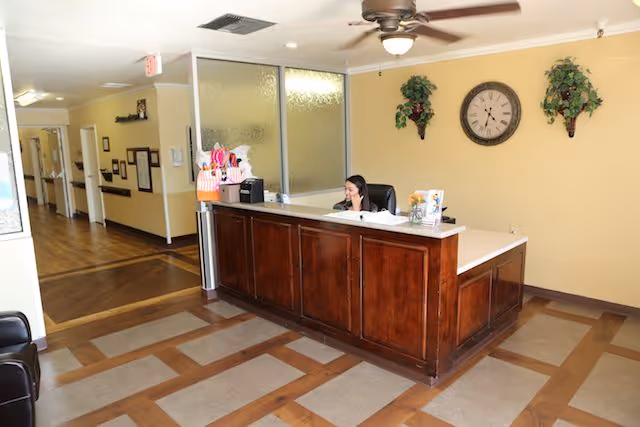 Reception area of a senior living facility with a wooden front desk where a woman is seated and talking on the phone. The room has a ceiling fan, a clock on the wall, two decorative plants, and a hallway leading to other rooms.