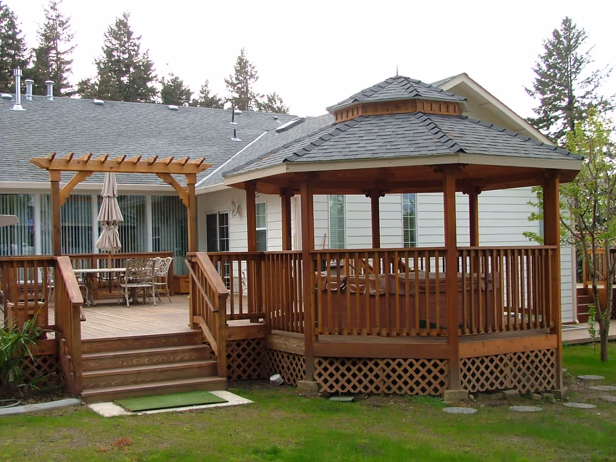 Outdoor wooden gazebo and deck attached to a white building with a gray roof. The deck has a wooden pergola with a closed umbrella and metal chairs and table. The area is surrounded by green grass and trees.